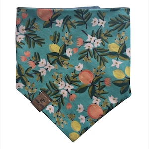 May include: A triangular pet bandana in a teal fabric featuring a repeating pattern of oranges, yellow lemons, white blossoms, and green leaves. A small brown rectangular leather-like tag is attached to the lower left corner with text.