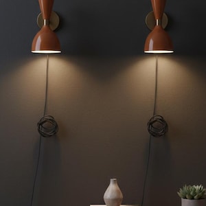 May include: Two matching orange wall sconces with brass detailing are mounted on a dark grey wall. A wooden cabinet below holds books, a vase, and a potted succulent. The sconces have a mid-century modern design.