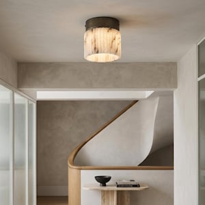 May include: A cylindrical flush mount ceiling light fixture featuring a dark bronze metal base and a translucent, vertically ribbed alabaster stone shade that emits a warm, diffused glow from the centre of the ceiling.