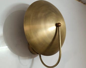 May include: A brass wall sconce with a large, rounded shade and a curved handle. The sconce is mounted on a white wall, casting a shadow. The design is minimalist and modern.