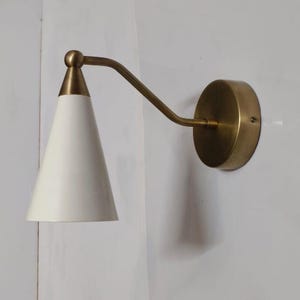 Pode incluir: Luminária de parede com abajur cônico branco e braço e placa de montagem na cor latão. A luminária tem um design minimalista, adequado para vários estilos de interiores. A sombra é inclinada para baixo.