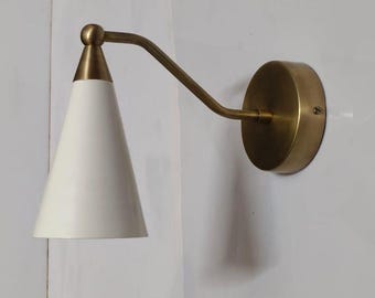 May include: A wall-mounted light fitting featuring a white, cone-shaped lampshade and a brass-coloured arm and mounting plate. The minimalist design is suitable for various interior styles. The shade is angled downwards.
