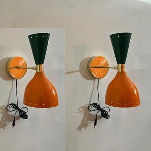 May include: Two mid-century modern wall sconces. Each lamp has a dark green conical shade, an orange, bell-shaped body, and a gold-coloured mounting plate. The lamps are wired with black cords and plugs.
