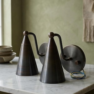 May include: Two bronze wall sconces with a conical shade and a circular backplate. The sconces have a curved arm and a small sphere at the top. The wires are blue, yellow, and white. The sconces are on a marble surface.