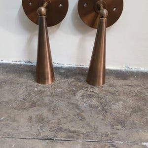 May include: Two bronze wall sconces. Each has a circular base and a conical shade. The sconces are mounted on a white wall and sit on a concrete floor. The sconces are a warm, metallic colour.