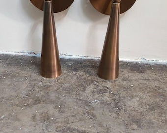 May include: Two bronze-colored wall sconces. Each sconce has a circular base with three screw holes and a conical shade. The sconces are mounted on a white wall and sit on a concrete floor.