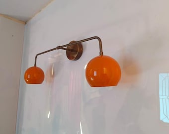 May include: A modern wall-mounted light fixture with two orange globe-shaped shades. The fixture has a bronze finish and is mounted on a white wall. The shades are a vibrant orange color.