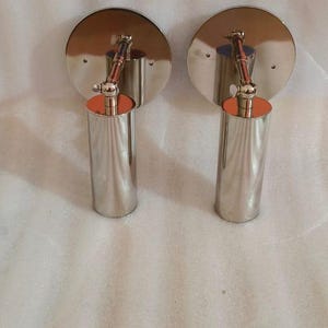 May include: Two modern wall sconces with a polished chrome finish. Each sconce features a circular backplate, an adjustable arm, and a cylindrical light fixture with an orange accent. The sconces are designed for indoor lighting.