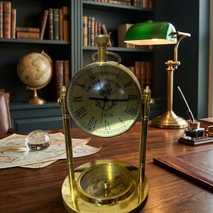 May include: A brass desk clock with a compass base, featuring a clock face with Roman numerals and the text "VICTORIA LONDON 1876". A green-shaded desk lamp and a globe are visible in the background, suggesting a vintage study or office setting.