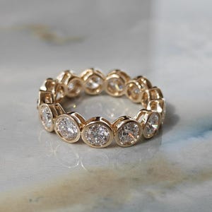 May include: A gold eternity band featuring a continuous row of round, brilliant-cut clear stones. Each stone is individually set in a polished gold bezel setting, creating a circular ring design with a uniform, metallic, and reflective appearance.