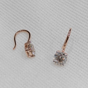 May include: A pair of rose gold drop earrings, each featuring a single round brilliant-cut clear gemstone set in a four-prong basket. The earrings are suspended from curved wire hooks, showcasing a polished metallic finish and faceted stones.