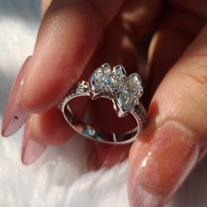 May include: A silver-toned ring featuring three marquise-cut clear stones arranged horizontally across the top. The band is thin and encrusted with small, sparkling clear stones along the sides, creating a bright and reflective appearance.