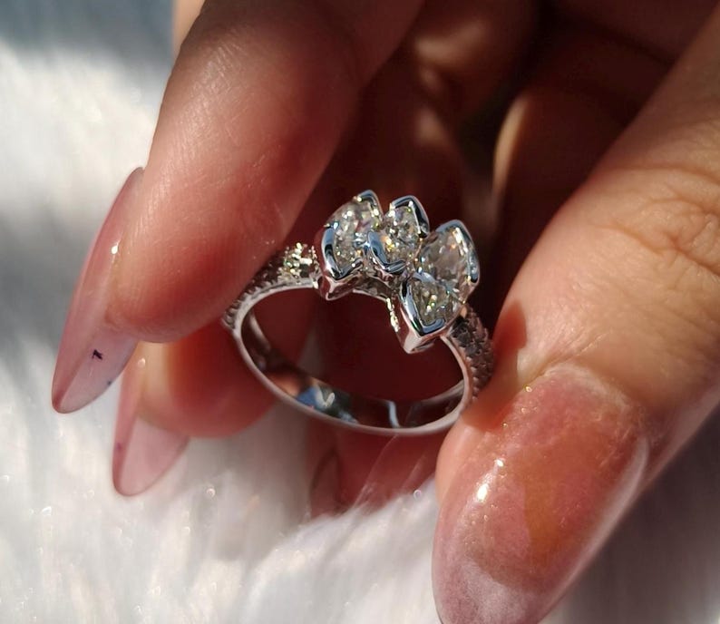 May include: A silver-toned ring featuring three marquise-cut clear stones arranged horizontally across the top. The band is thin and encrusted with small, sparkling clear stones along the sides, creating a bright and reflective appearance.