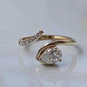 Op de afbeelding: Een open gouden ring met een peervormige diamant in een gladde gouden zetting aan het ene uiteinde en een rij kleine ronde pavé-gezette diamanten langs het andere taps toelopende uiteinde, met een gepolijste afwerking en heldere stenen.