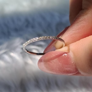 Può includere: Un delicato anello d'argento con una fascia di piccoli diamanti scintillanti. L'anello è tenuto tra le dita, mettendo in risalto i suoi dettagli raffinati e la brillantezza delle pietre preziose. L'anello è un gioiello.