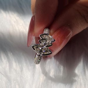 May include: A silver-toned ring featuring two marquise-cut clear stones arranged in a bow-like shape, set on a band encrusted with small, sparkling pav&eacute; stones. The metal band has a polished finish and reflects light across its faceted surfaces.