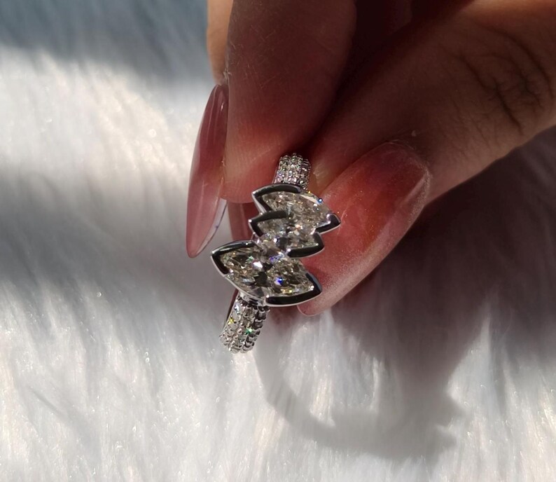 May include: A silver-toned ring featuring two marquise-cut clear stones arranged in a bow-like shape, set on a band encrusted with small, sparkling pav&eacute; stones. The metal band has a polished finish and reflects light across its faceted surfaces.