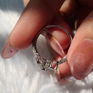 May include: A silver-toned ring featuring a central bow-shaped design adorned with small, clear, sparkling stones. The band is thin and also encrusted with a series of tiny, clear, reflective stones along its outer surface, creating a continuous shimmering effect.
