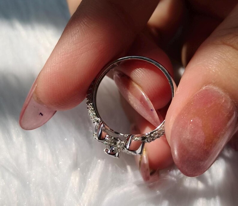 May include: A silver-toned ring featuring a central bow-shaped design adorned with small, clear, sparkling stones. The band is thin and also encrusted with a series of tiny, clear, reflective stones along its outer surface, creating a continuous shimmering effect.