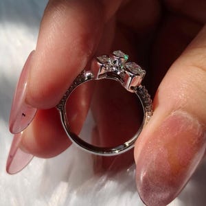 May include: A silver-toned ring featuring a cluster of three clear, faceted stones set at the top. The band is metallic and smooth on the interior, with a textured, pav&eacute;-style surface along the outer edges leading up to the central stone setting.