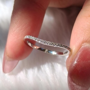 Op de afbeelding: Een gebogen, zilverkleurige ring met een rij kleine, sprankelende edelstenen. De ring wordt tussen vingers gehouden, waardoor het delicate ontwerp en het reflecterende oppervlak worden benadrukt. De ring lijkt gemaakt van glanzend metaal, mogelijk platina of witgoud.