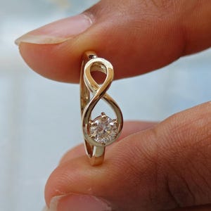 Può includere: Un anello in oro caratterizzato da un design a simbolo di infinito sulla fascia. L'anello dell'infinito è impreziosito da una singola pietra preziosa rotonda, chiara e sfaccettata, incastonata saldamente nella curva inferiore del simbolo.