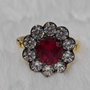 Peut inclure: Une bague en or avec une grande pierre centrale ronde rouge foncé entourée d'un halo de dix pierres plus petites, rondes, claires et facettées. La pierre centrale est sertie dans une monture en métal sombre et l'anneau est en or jaune.