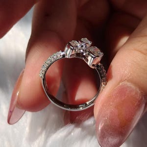 May include: A silver-toned ring featuring a cluster of three clear, faceted stones arranged in a floral-like pattern at the top. The band is slender and encrusted with a row of small, sparkling clear stones along the sides of the shank.