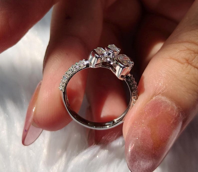 May include: A silver-toned ring featuring a cluster of three clear, faceted stones arranged in a floral-like pattern at the top. The band is slender and encrusted with a row of small, sparkling clear stones along the sides of the shank.