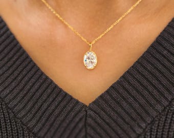 May include: A gold-colored necklace with a pendant featuring a large, oval-shaped, clear gemstone. The necklace is worn against a dark gray v-neck sweater, creating a contrast that highlights the jewelry.