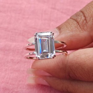 May include: A silver-coloured ring featuring a large, rectangular emerald-cut clear gemstone set in a prong setting. The band is split into two thin, parallel silver-coloured metal wires that connect at the base of the central stone setting.