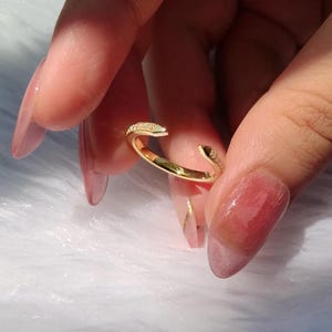 Könnte beinhalten: Ein goldfarbener offener Ring mit kleinen, klaren Edelsteinen an den oberen Kanten. Der Ring wird vor einem weichen, weißen Hintergrund präsentiert. Die Nahaufnahme zeigt die Details des Rings und das Licht, das sich in den Edelsteinen spiegelt.