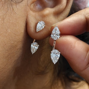 May include: A pair of silver-toned earrings featuring a double pear-shaped design. Each earring consists of a smaller pear-shaped stone at the top and a larger pear-shaped stone suspended below by a thin metal bar, creating a drop effect.