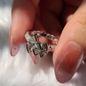 May include: A silver-toned ring featuring three prominent marquise-cut clear stones arranged horizontally in the center, flanked by bands encrusted with small, shimmering pav&eacute;-set stones along the sides of the metal shank.