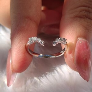 May include: A silver ring with a unique open design, featuring a row of small, sparkling diamonds on each end. The ring is held up against a soft, white background, showcasing its elegant and modern style. The diamonds catch the light, creating a brilliant display.