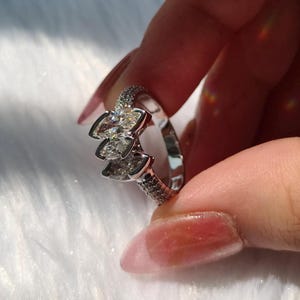 May include: A silver-toned ring featuring three marquise-cut clear stones arranged in a cluster at the center. The band is polished and accented with small pav&eacute;-set clear stones along the sides, creating a reflective and metallic appearance.