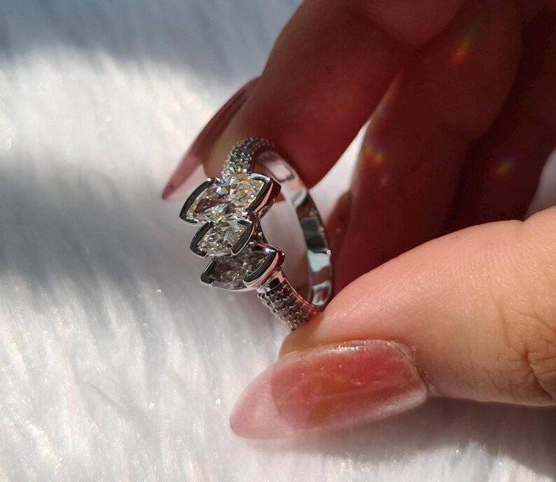 May include: A silver-toned ring featuring three marquise-cut clear stones arranged in a cluster at the center. The band is polished and accented with small pav&eacute;-set clear stones along the sides, creating a reflective and metallic appearance.