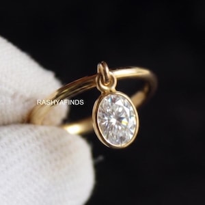 May include: A gold ring featuring a thin, smooth band and a single oval-shaped, clear, faceted stone set in a bezel setting that hangs from a small loop. The text RASHYAFINDS is printed on the side of the band. The ring has a polished finish.