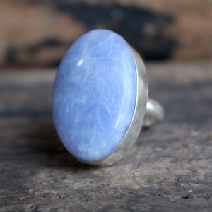 May include: Oval-shaped blue gemstone ring with a silver band. The light blue stone is set in a silver bezel, creating a simple yet elegant design. The ring is displayed on a wooden surface.