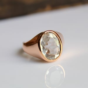 Puede incluir: Un anillo de oro rosa con una gema grande, de talla ovalada, de color verde p&aacute;lido. El anillo est&aacute; engastado en una banda lisa y pulida, y la gema refleja la luz. El anillo se exhibe sobre una superficie blanca.