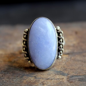 May include: Oval-shaped silver ring featuring a large, light blue gemstone. The ring has a detailed silver band with decorative elements on the sides. The gemstone is smooth and polished, set in a classic bezel setting.