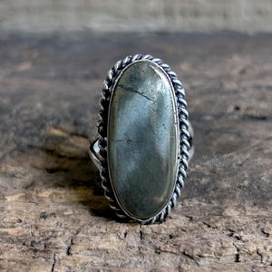 May include: A silver ring with an oval, gray-green stone. The stone is set in a silver band with a twisted rope design around the edge. The ring is displayed on a textured, brown wooden surface.