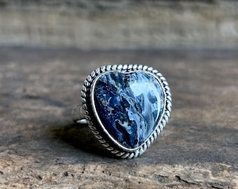 Peut inclure: Bague en argent ornée d'une pierre en forme de cœur, bleue et blanche. La pierre est sertie dans un cadre argenté torsadé. Posée sur une surface en bois rustique.