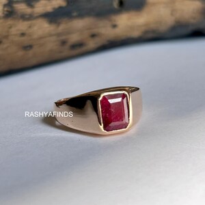 Op de afbeelding: Een goudkleurige ring met een rechthoekige, dieprode edelsteen. De ring heeft een eenvoudig, klassiek ontwerp met een gepolijste afwerking. De tekst "RASHYAFINDS" is zichtbaar op de ring.
