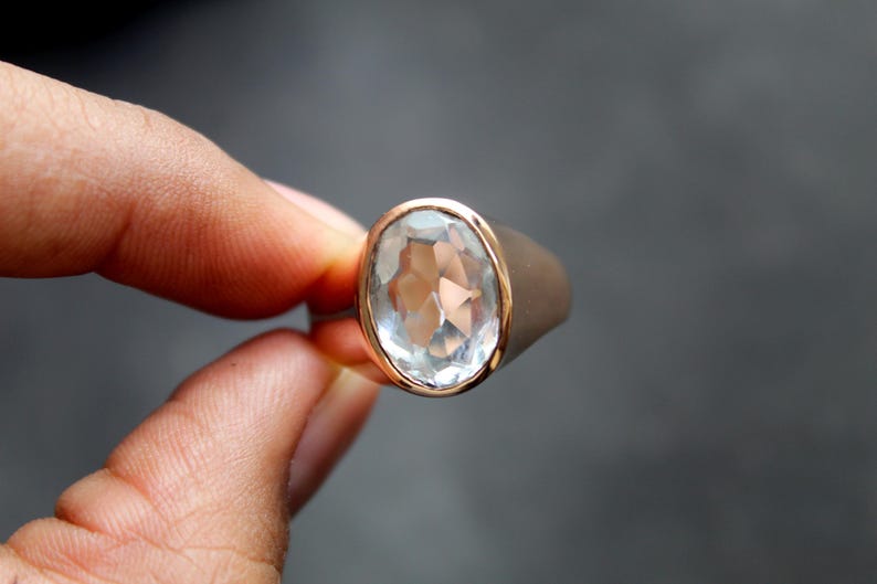 Puede incluir: Un anillo plateado con una gema grande, ovalada, facetada y transparente. La gema est&aacute; engastada en un bisel dorado. El anillo se sostiene entre dos dedos sobre un fondo gris oscuro. El anillo es una joya.