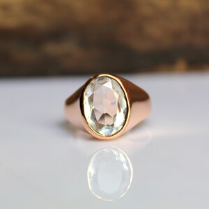 Puede incluir: Un anillo de oro rosa con una piedra preciosa ovalada y clara. El anillo tiene una banda lisa y pulida. La piedra preciosa refleja la luz, y el anillo se muestra sobre una superficie blanca con un fondo borroso.