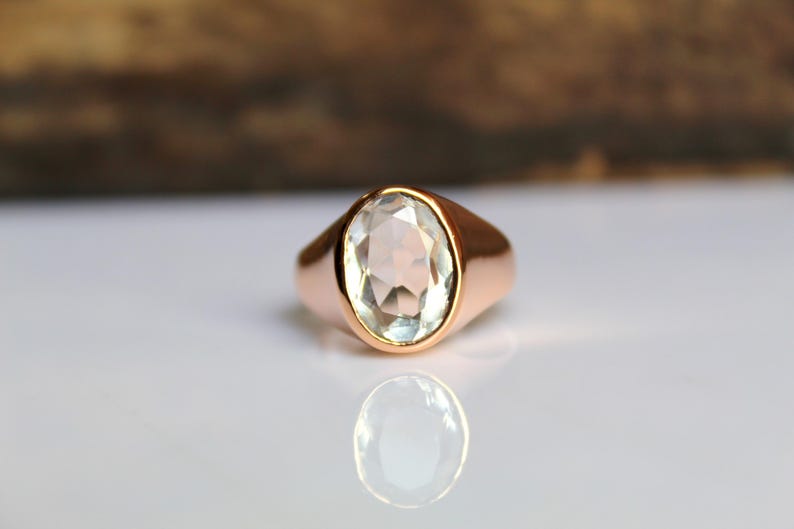 Puede incluir: Un anillo de oro rosa con una piedra preciosa ovalada y clara. El anillo tiene una banda lisa y pulida. La piedra preciosa refleja la luz, y el anillo se muestra sobre una superficie blanca con un fondo borroso.