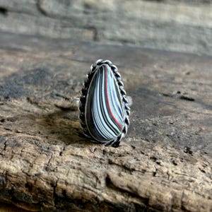 May include: A silver ring with a teardrop-shaped stone. The stone has vertical stripes in black, white, red, and blue. The ring has a twisted rope-like border.