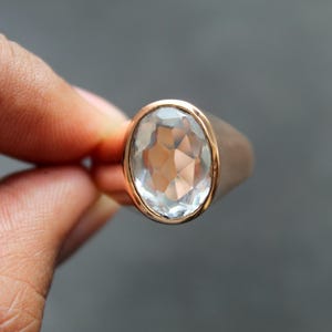 Puede incluir: Un anillo de oro rosa con una gema clara y facetada de gran tama&ntilde;o. La piedra de forma ovalada est&aacute; engastada en un bisel dorado. El anillo se sostiene entre dos dedos sobre un fondo gris. Es una joya.