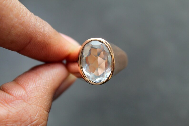 Puede incluir: Un anillo de oro rosa con una gema clara y facetada de gran tama&ntilde;o. La piedra de forma ovalada est&aacute; engastada en un bisel dorado. El anillo se sostiene entre dos dedos sobre un fondo gris. Es una joya.